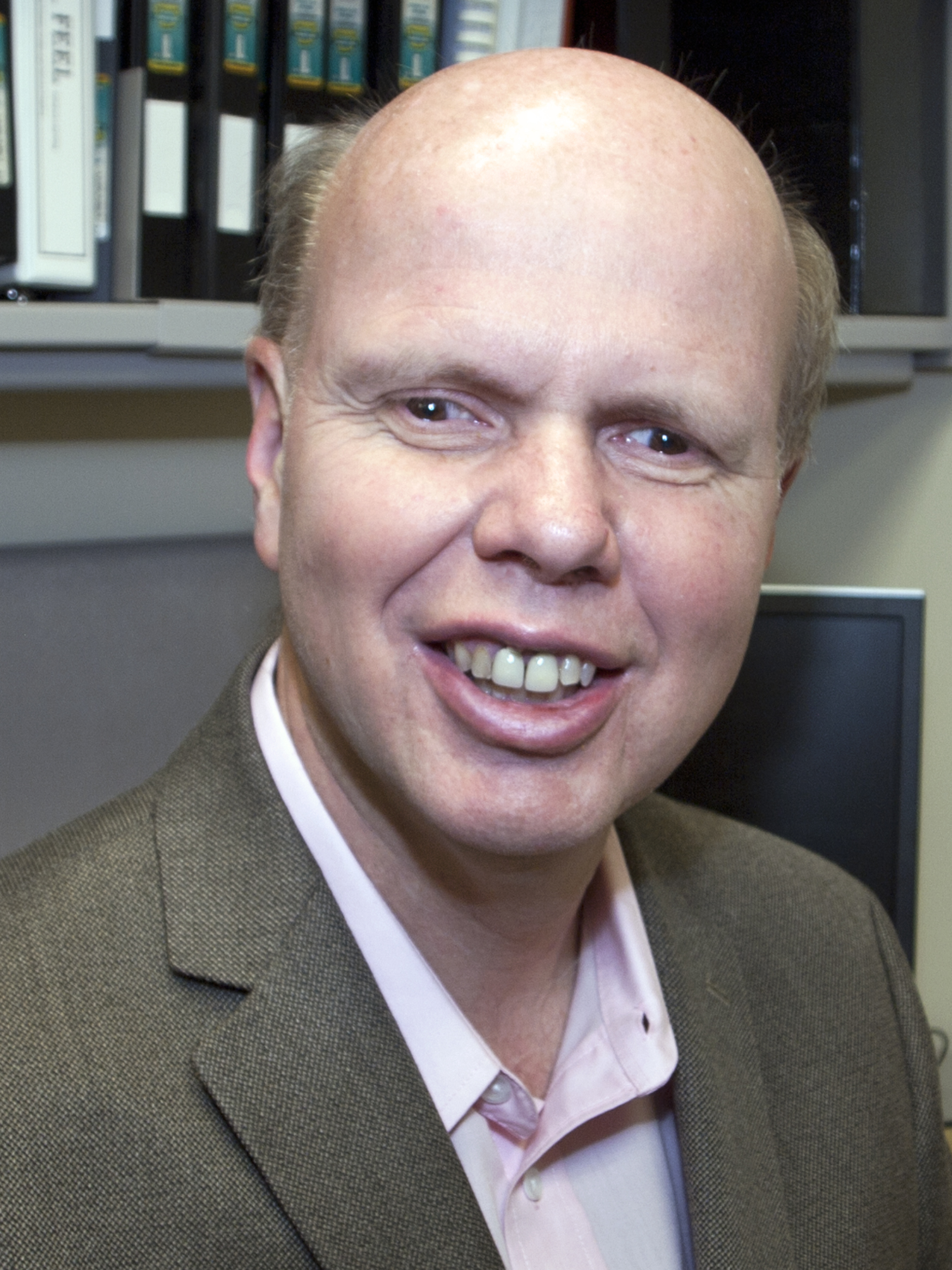 A professional image of Mike Hoenig. He is smiling and wearing a brown suit jacket with a pastel collared shirt. 
