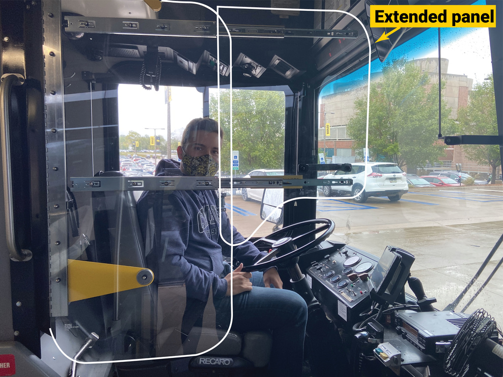 man sitting in drivers seat behind transparent driver barrier panel with driver panel extended to cover more of drivers area - gold box with text "panel extended' to aid in seeing transparent panel