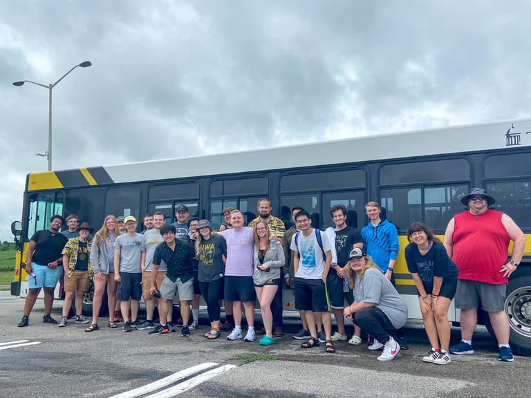 group photo of many people standing in front of gold and black transit bus on sunny, warm day