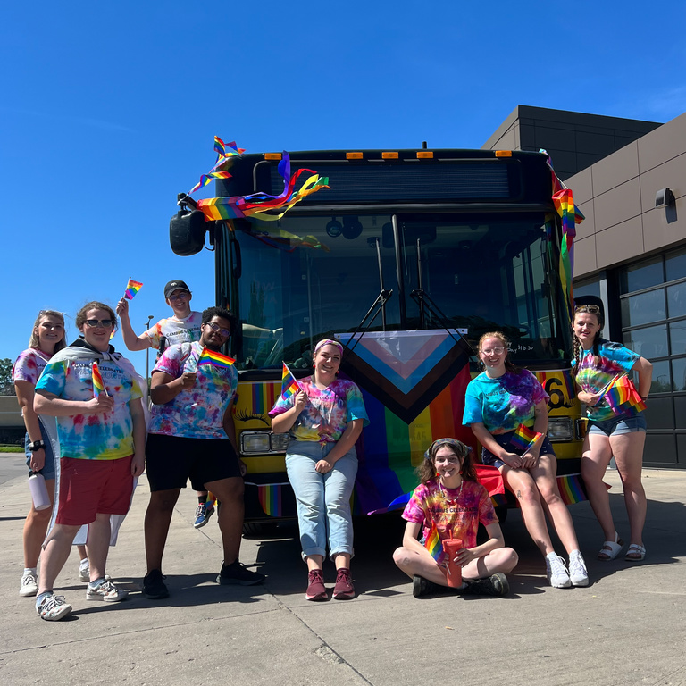 Student employees smiling outside a bus decorated for pride