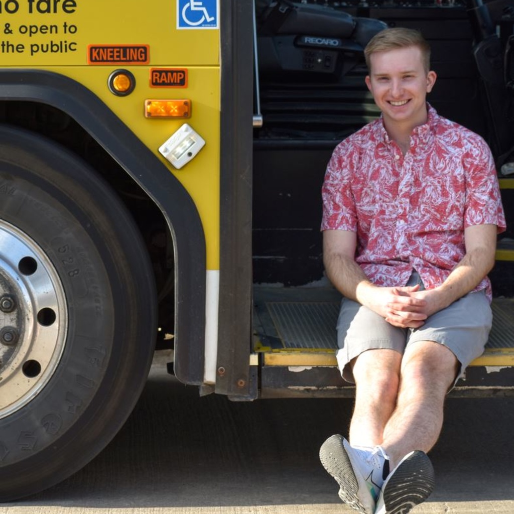 Person seated in bus doorway wearing khaki shorts and a red aloha shirt