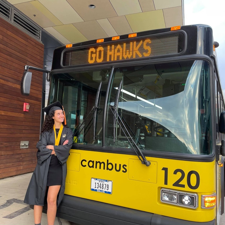 CAMBUS student employee standing next to a bus, wearing a graduation cap and gown
