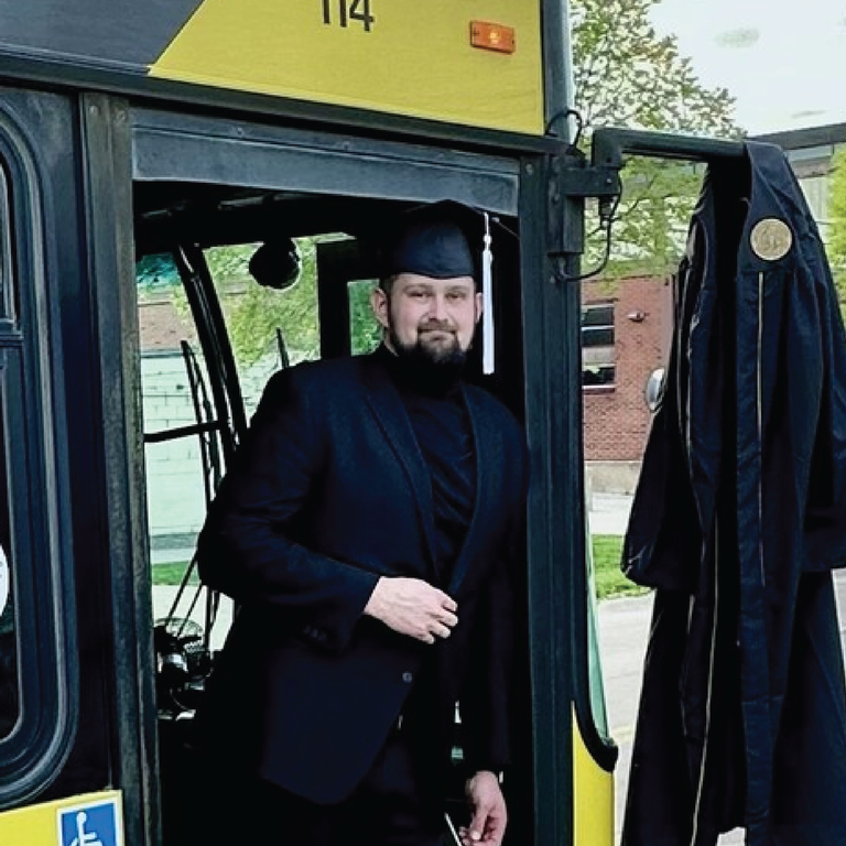 Ben poses with a cap and gown in the entry way of a bus