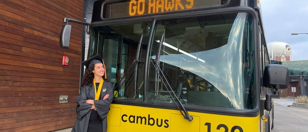 CAMBUS student employee standing next to a bus, wearing a graduation cap and gown