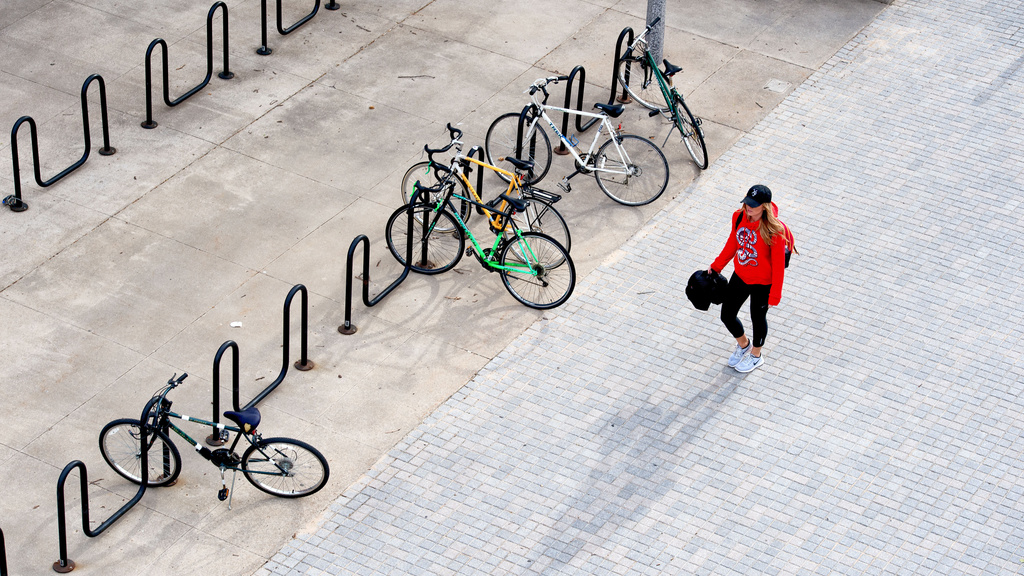 person wearing black ball cap and red sweatshirt walks past two rows of bicycle racks with bikes parked