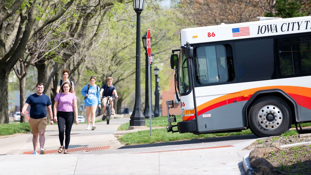 persons walking and biking on campus side walk near intersection with Iowa City Transit bus at stop sign